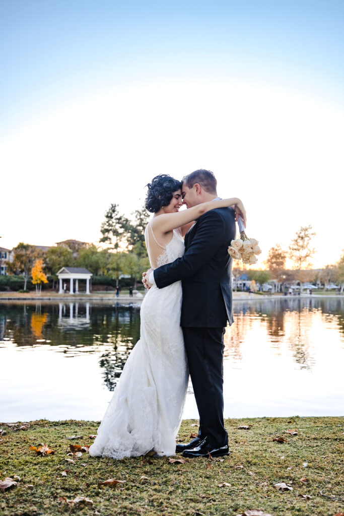 Bride and groom embrace beside peaceful lake at golden hour after Catholic wedding