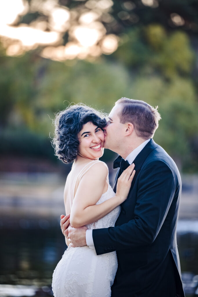 Bride smiles at camera as groom holds her close beside Murrieta lakeside