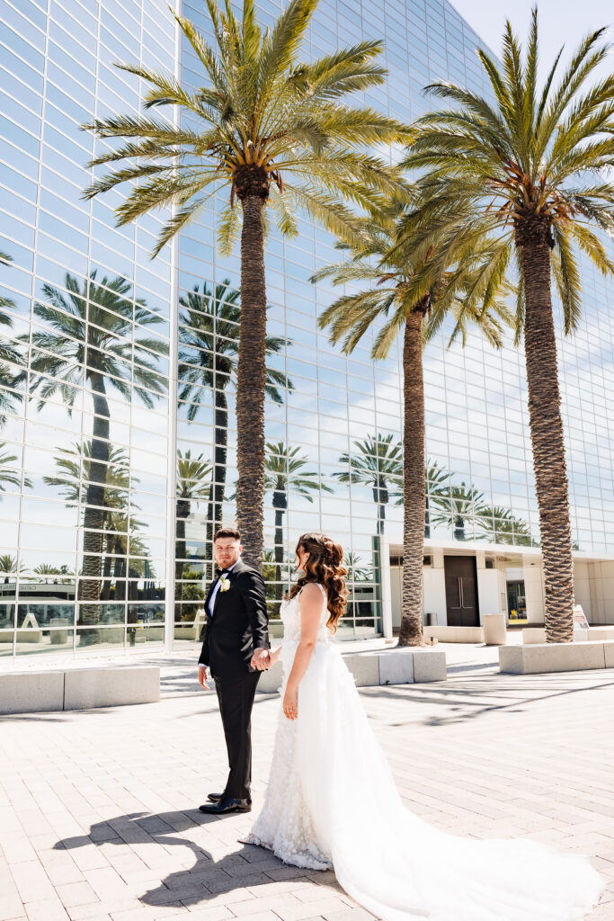 Wide angle wedding portrait of bride and groom standing on the plaza at Christ Cathedral Garden Grove surrounded by tall palm trees and reflective glass architecture.
