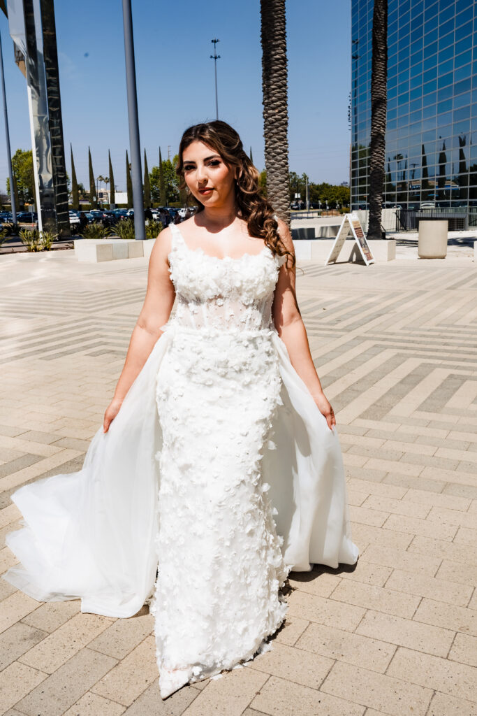 Bride standing on the plaza at Christ Cathedral in Garden Grove wearing a fitted 3D floral lace wedding dress with a detachable train.