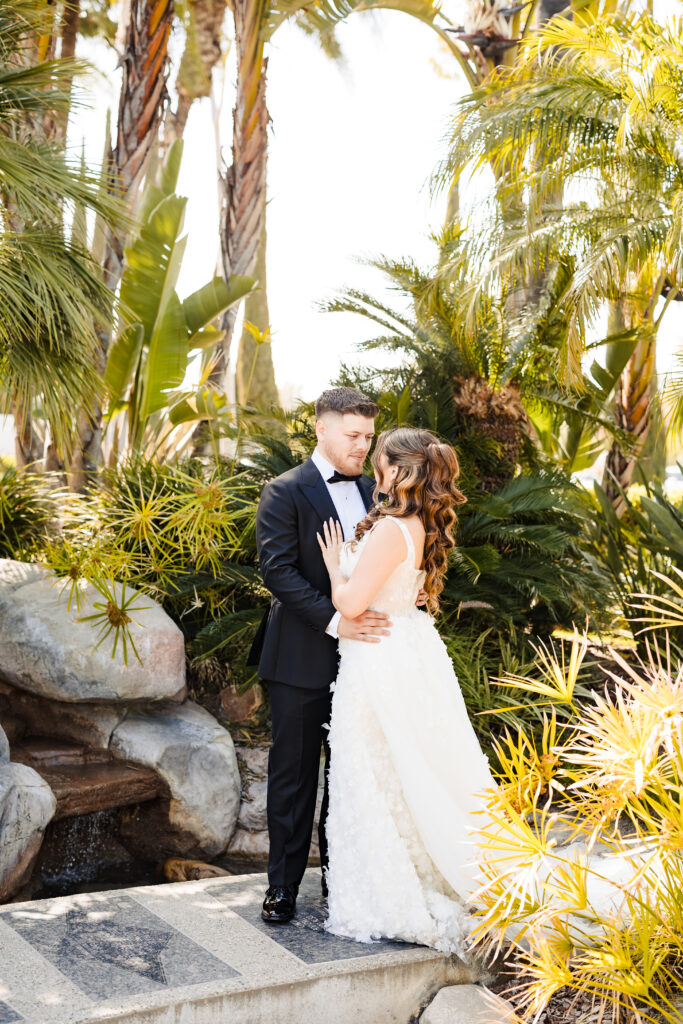 Bride and groom embracing in the lush tropical gardens at Christ Cathedral in Garden Grove, surrounded by palm trees and greenery.